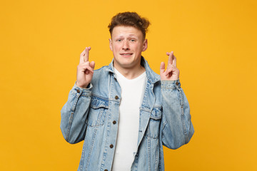 Young man in denim casual clothes waiting for special moment, keeping fingers crossed, making wish isolated on yellow orange background. People sincere emotions lifestyle concept. Mock up copy space.