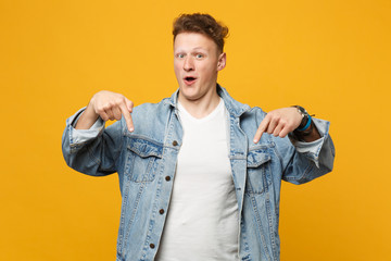 Excited young man in denim casual clothes keeping mouth open, pointing index fingers down isolated...
