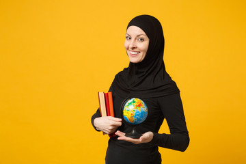 Smiling young arabian muslim student girl in hijab black clothes hold in hands globe, books isolated on yellow wall background, studio portrait. People religious lifestyle concept. Mock up copy space.