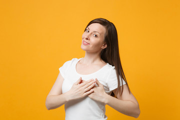 Portrait of pleasant young woman in white casual clothes looking camera keeping hands on chest, heart isolated on yellow orange wall background in studio. People lifestyle concept. Mock up copy space.