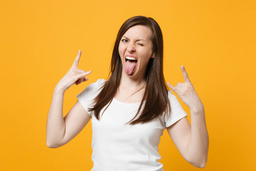 Crazy blinking young woman in white casual clothes depicting heavy metal rock sign, horns up gesture showing tongue isolated on yellow orange background. People lifestyle concept. Mock up copy space.