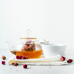 White teacups and transparent teapot with herbal tea of dried pink roses buds on white background with copy space. Brewing and Drinking tea.