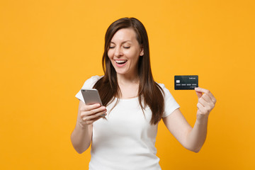 Portrait of pretty young woman in white casual clothes using mobile phone, holding credit bank card isolated on yellow orange wall background in studio. People lifestyle concept. Mock up copy space.