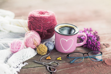  Women's hobby. Crochet and knitting. Working space. Bouquet of lilac in a cup, balls of yarn, needles, scissors, knitwear, crochet hooks on the wood table  in the cozy home. 
