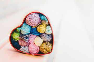 Heart-shaped box with knitted multi-colored skeins of yarn on wooden background in the garden on spring day. Crochet and knitting. Women's working space. 