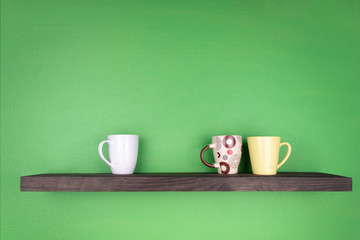 a dark color shelf with texturing wood is installed on a green wall; there are three different color mugs on the shelf