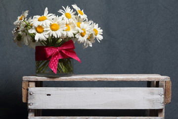 A bouquet of field daisies in a glass vase. On the  wooden box. Genus of perennial flowering plants of the Matricária family (Matricária).