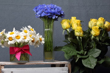 A bouquet of field daisies, cornflowers and yellow roses in glass vases. On a gray background.