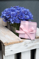 Bouquet of cornflowers in a glass vase. Standing on a wooden box. Next gift, packaged in a box and tied with a bow. On a gray background.
