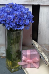 Bouquet of cornflowers in a glass vase. Stands near the wooden box. Near photo album, a gift, packed in a box and tied with a bow. On a gray background.