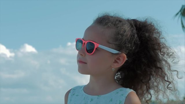 Close-up Portrait of a Beautiful Little Girl in Pink Glasses, Cute Smiling, Looking at the Blue Sky. Concept: Children, Childhood, Summer, Baby, Toddler.