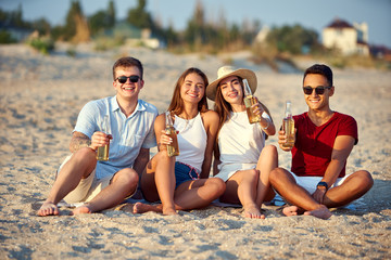 Group of friends having fun enjoying a beverage and relaxing on the beach at sunset in slow motion. Young men and women drink beer sitting on a sand in the warm summer evening.