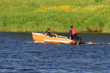 Two men in life jackets floating in a small orange fishing outboard motor boat along the shore with green grass, rear side view