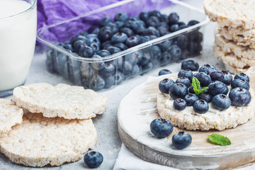Healthy organic rice cakes with ricotta and fresh blueberries and glass of milk on light stone kitchen background. Top view. Plastic tray of strawberries