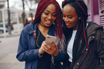 two beautiful and stylish dark-skinned girls with long hair standing in a autumn city near purple wall and using the phone