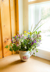 Vintage style flower bouquet made of wild flowers found in forest and meadow, standing in old cream jug on window sill, summers in grandma`s house concept. Northern Europe.