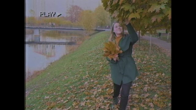 Young woman walks in the autumn park. Cheerful woman with blond hair in a green coat walks on the yellow leaves. Girl with a bouquet of leaves in her hands. Old video. Home video archive. Family video