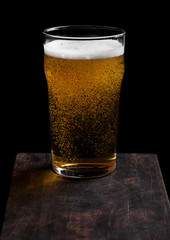 Glass of lager beer with foam and bubbles on vintage wooden board on black background.