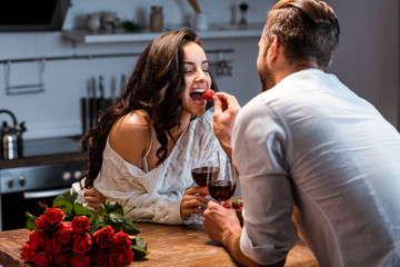couple with glasses of red wine at wooden table with bouquet of roses, man feeding woman with strawberry