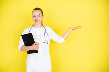 Young female blonde doctor with stethoscope on her neck and tablet points her finger on a space near her. Place for text