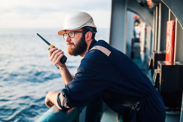 Deck Officer on deck of offshore vessel or ship , wearing PPE personal protective equipment. He holds VHF walkie-talkie radio in hands. Dream work at sea - obrazy, fototapety, plakaty