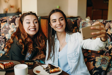 Two amazing young girlfriends doing a funny selfie while sitting on a coffee shop having fun together.