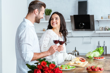 happy man clinking glasses of red wine with smiling woman at kitchen