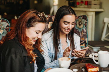 Portrait of two beautiful young woman sitting in a coffee shop and looking at their smartphone smiling navigating on internet.