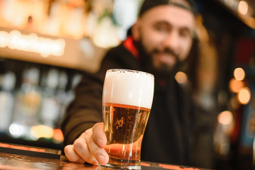 Bearded and smiling barman gives beer. Cool and courageous bar. A happy barman holds a beer in his hand.