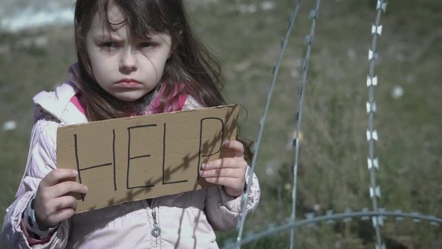 A child in a refugee camp asks for help. Sad little girl behind barbed wire.