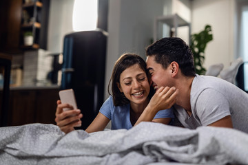 Affectionate couple using smart phone and taking selfie in bedroom.