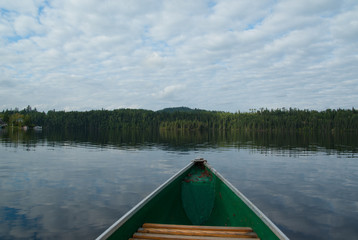 Fototapeta premium Picture of a canoe on a calm lake with fluffy clouds reflecting in the water. Rolling hills and green forest with cottages on the horizon. Very peaceful and propitious to meditation.