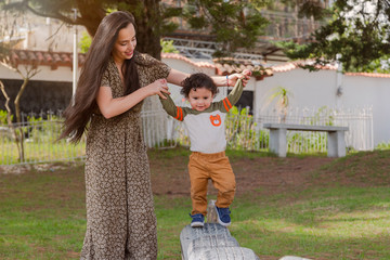 Latina mom taking her little son by the hands and helping him walk in the park