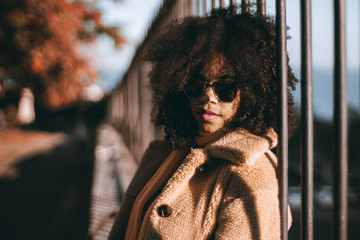 The portrait of a young African-American female leaning against an iron fence outdoors in the park; beautiful curly-hair biracial girl in sunglasses in a warm coat outdoors