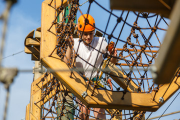 Fototapeta premium Child in forest adventure park. Kid in orange helmet and white t shirt climbs on high rope trail. Agility skills and climbing outdoor amusement center for children. young boy plays outdoors