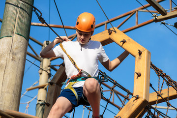 Child in forest adventure park. Kid in orange helmet and white t shirt climbs on high rope trail. Agility skills and climbing outdoor amusement center for children. young boy plays outdoors