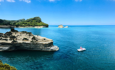 Wide seascape scene of Mediterranean Sea ( Corfu, Greece). In foreground is pedal boat with tourists, next to boat is cliff with specific structure of stone and in the background is green vegetation.
