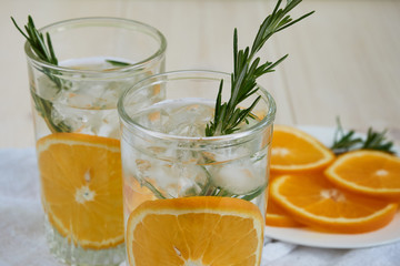 Two glasses of cool refreshing drink with ice, orange and rosemary on a white napkin, close-up
