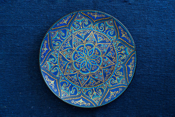 Decorative ceramic plate with blue and golden colors, painted plate on the background of blue fabric, closeup. Decorative porcelain plate painted with acrylic paints, handwork