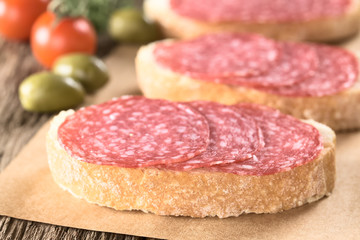 Salami slices on ciabatta bread with green olives and cherry tomato in the back (Selective Focus, Focus on the front of salami slices on first sandwich)