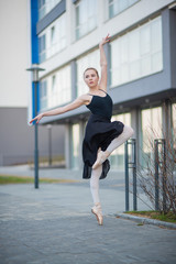 Ballerina in a tutu posing against the backdrop of a residential building. Beautiful young woman in black dress and pointe shoes dancing ballet outside.