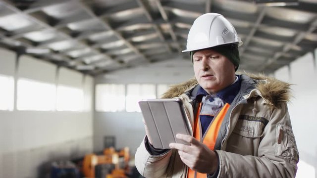 man in a hard hat and overalls is standing in the middle of the hangar and cheking tools with his tablet at the camera