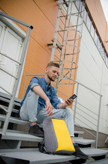 young man in jeans and denim shirt is sitting on stairs of industrial building with smartphone in his hand while waiting
