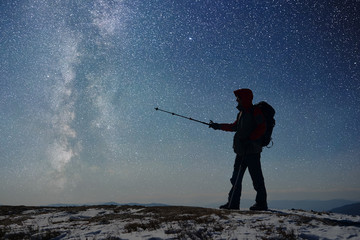 Silhouette of tourist pointing by trekking stick on Milky Way. Hiker with backpack standing on snow-covered top of mountains. Hiking in darkness. Starry sky, Milky Way on background. General side view