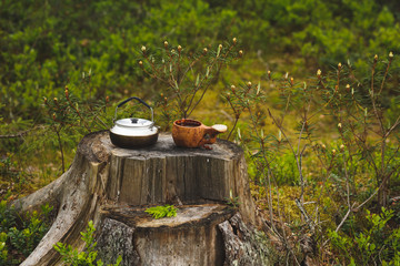 Obraz premium Wooden kuksa with coffee and kettle on the stump in focus. The forest background is blurred