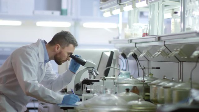 Tracking left of male researcher in medical gown standing at desktop in laboratory, using microscope and then making notes