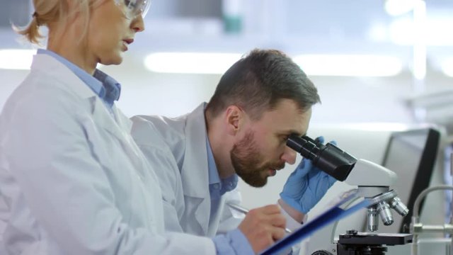 Tilt down of male and female researchers in medical gowns standing at desktop in lab. Man using microscope and woman writing down results into special form