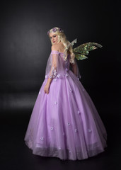  full length portrait of a blonde girl wearing a fantasy fairy inspired costume,  long purple ball gown with fairy wings,   standing pose with back to the camera on a dark studio background.