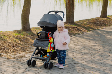 A little girl is rolling baby carriage in the park. Child in the park playing with pram.
