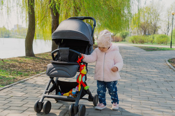 A little girl is rolling baby carriage in the park. Child in the park playing with pram.
